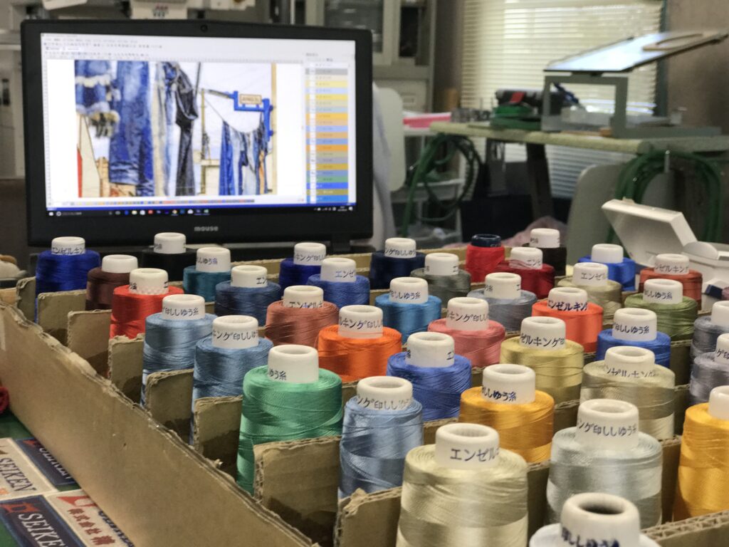A scene from a photo embroidery workspace, showing colorful spools of thread and a computer screen displaying embroidery software with a reference image.