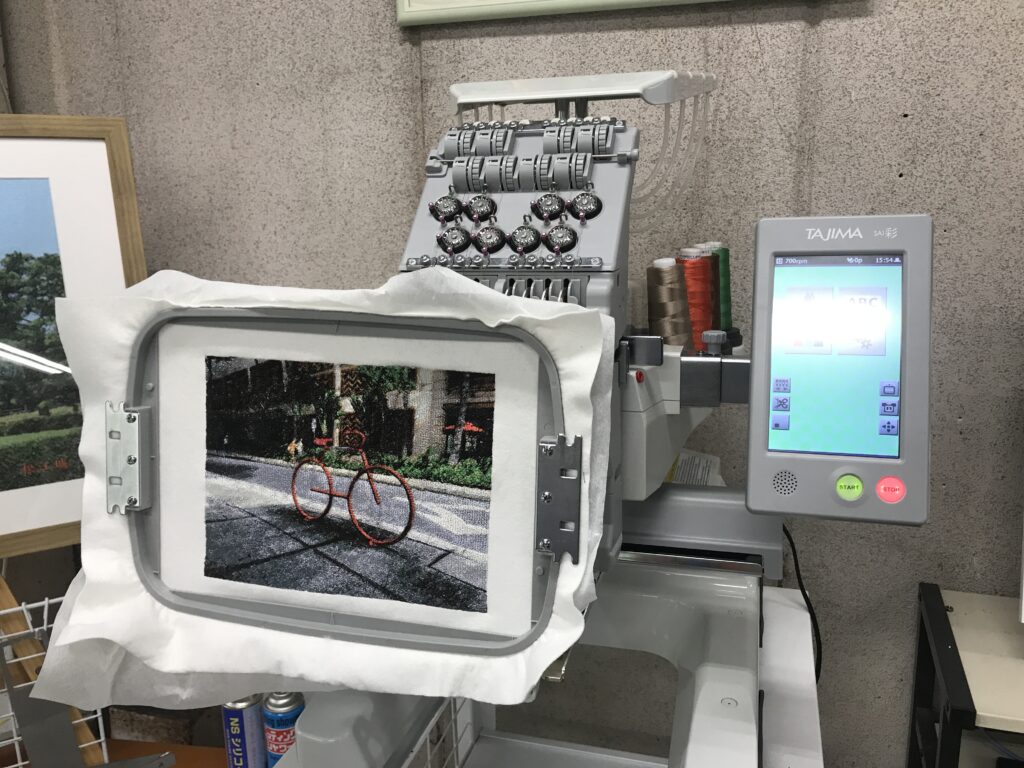 A Tajima embroidery machine working on a photo embroidery piece depicting a red bicycle in an urban scene. The work is shown mid-production in an embroidery hoop.