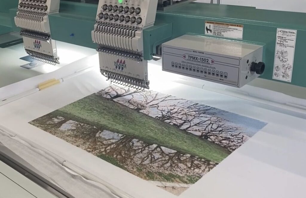 大型タジマ刺繍機で、背割堤の桜並木をモチーフにしたフォト刺繍作品を縫製している様子。緻密なステッチで春の風景を表現。