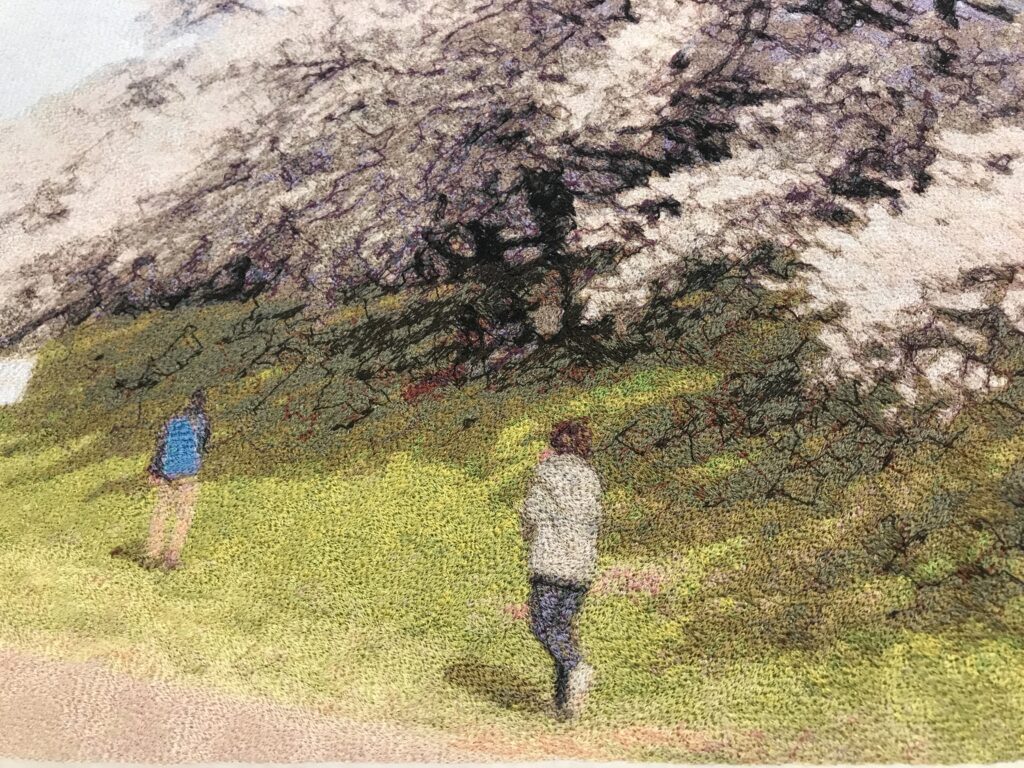 Close-up of the photo embroidery artwork “Cherry Blossoms at Sewaritei.” Depicts detailed stitching of people walking under blooming cherry trees and sunlight filtering onto the grass.