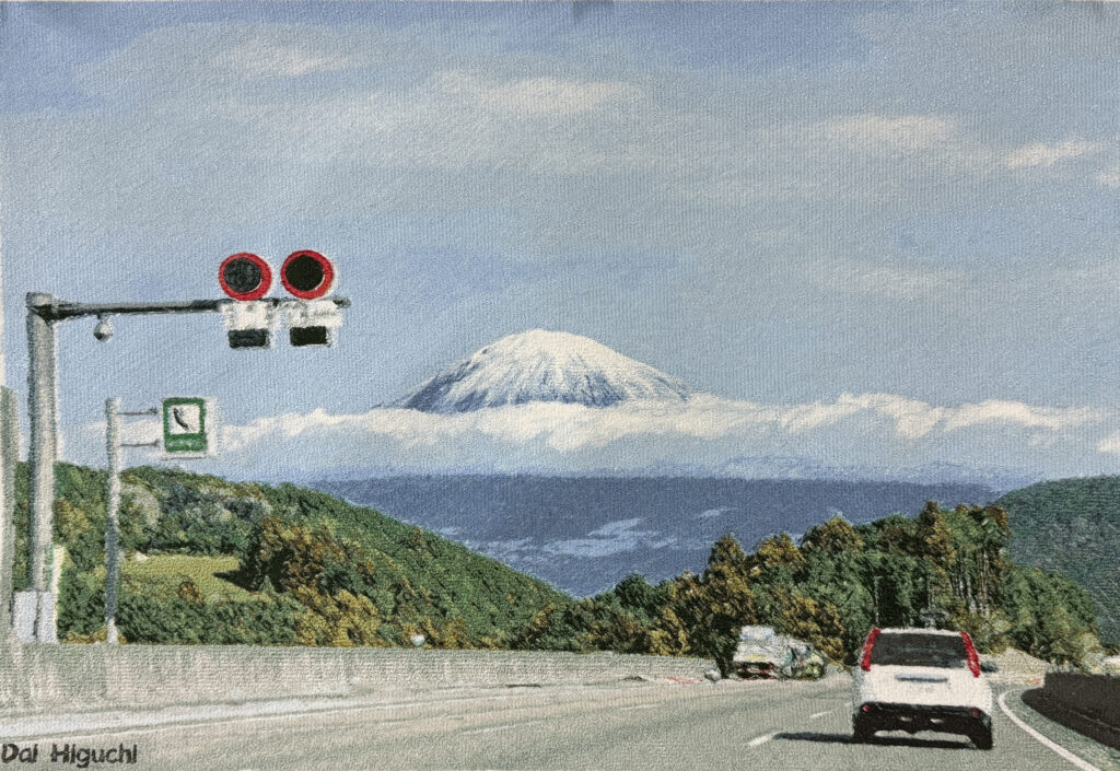 「高速道路を走る車越しに、雲をまとった富士山が見える情景を精密なフォト刺繍で表現した作品。前景には道路標識、周囲には緑の山並みが広がり、遠景の富士山が柔らかな糸の光沢で描かれている。」